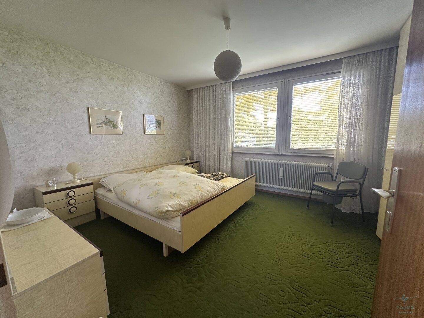 Schlafzimmer mit grünem Teppichboden, Doppelbett und Fenster mit Jalousien, bietet eine ruhige Atmosphäre.