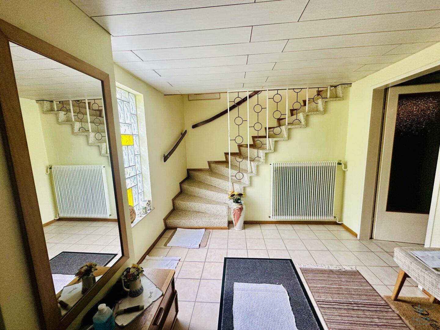 Heller Eingangsbereich mit Fliesenboden, großem Spiegel und einer geschwungenen Treppe zum Obergeschoss.