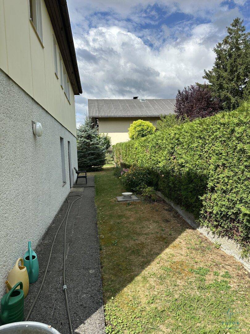 Seitenansicht des Hauses mit schmalem Weg entlang der Fassade und angrenzendem Gartenbereich.