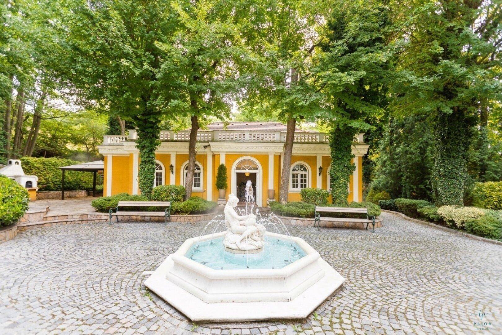 Elegantes Nebengebäude mit Springbrunnen und gepflastertem Hof, umgeben von hohen Bäumen.