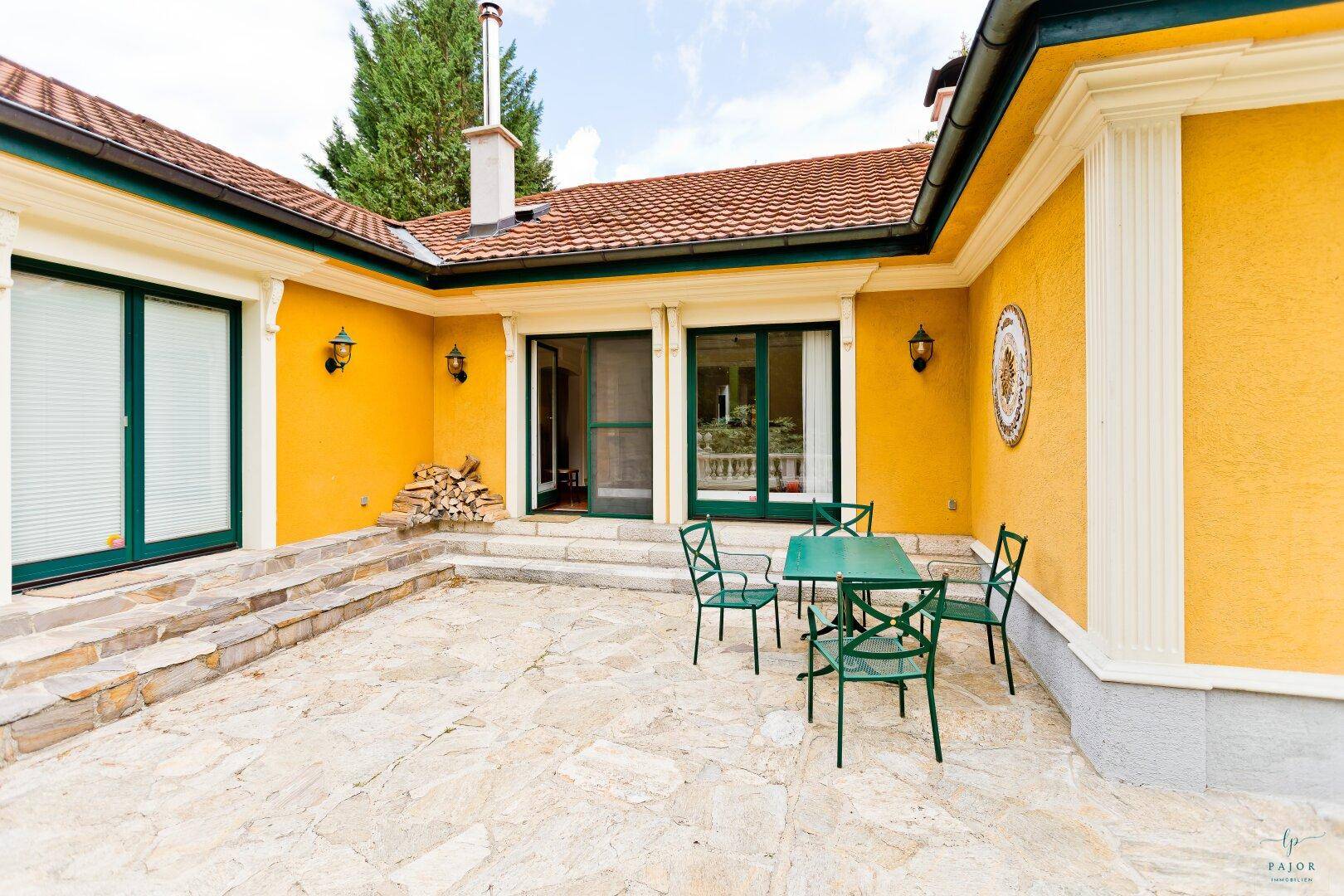 Sonniger Terrassenbereich mit Steinfliesen, grünen Gartenmöbeln und Holzstapel an der Hauswand.