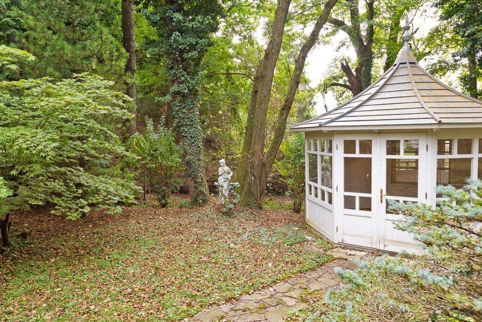 Idyllischer Gartenbereich mit weißem Pavillon und klassischer Statue unter hohen Bäumen.