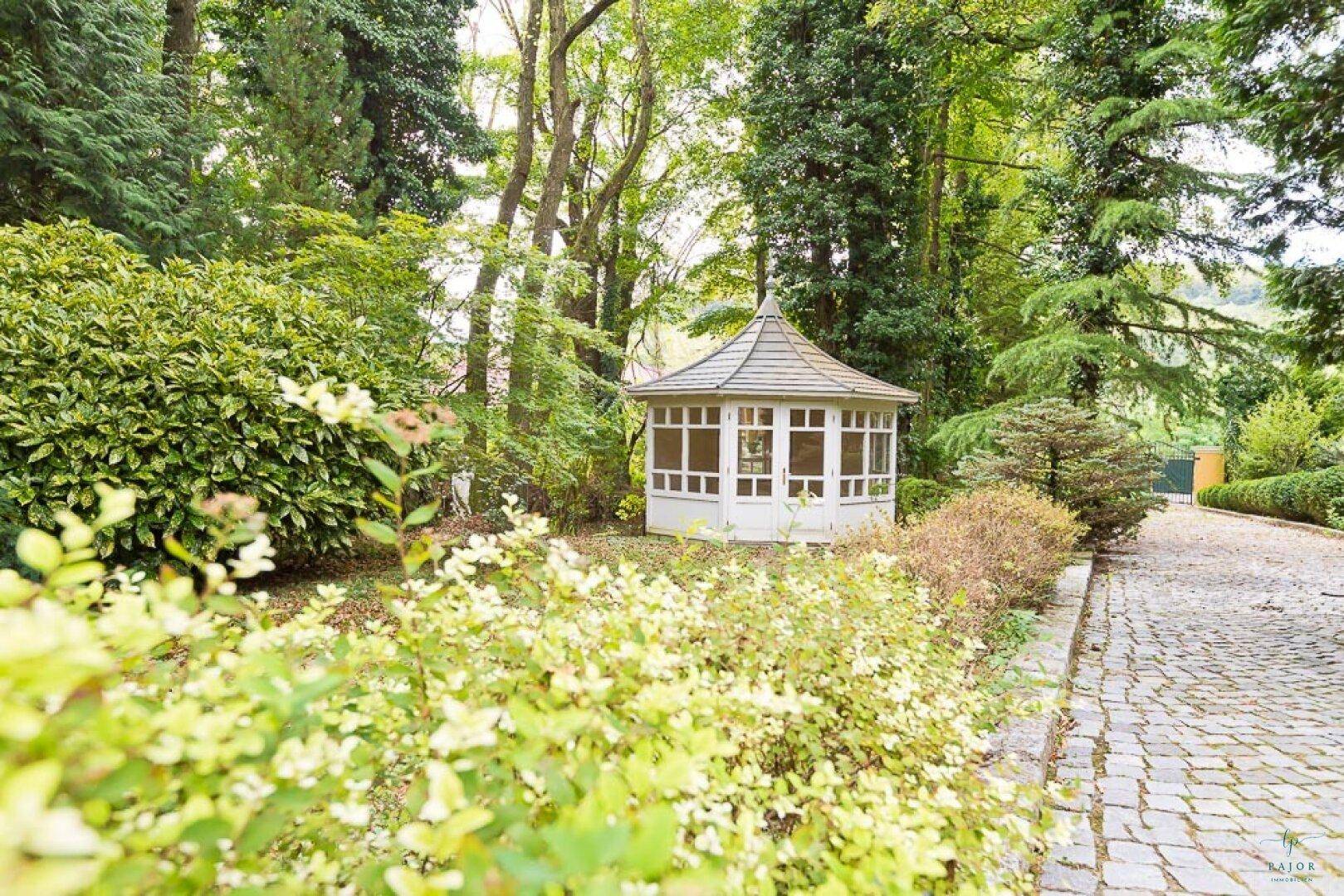 Malerischer Gartenweg führt zu einem charmanten weißen Pavillon, umgeben von Bäumen und Sträuchern.