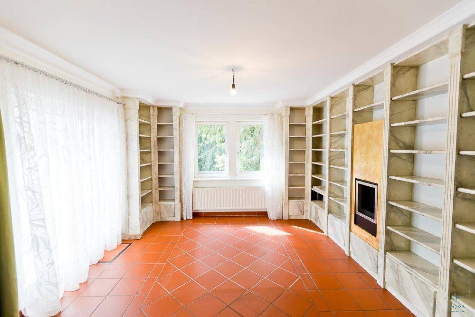 Raum mit raumhohen Bücherregalen, Terrakottafliesen und großen Fenstern.