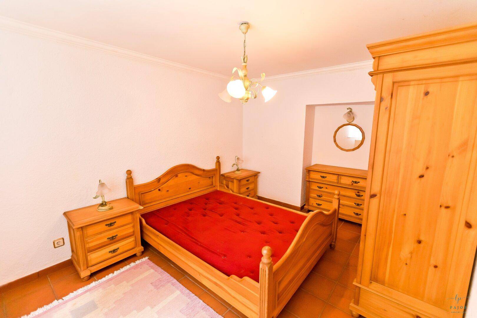 Schlafzimmer mit traditionellen Holzmöbeln, Terrakottafliesen und roter Bettdecke.