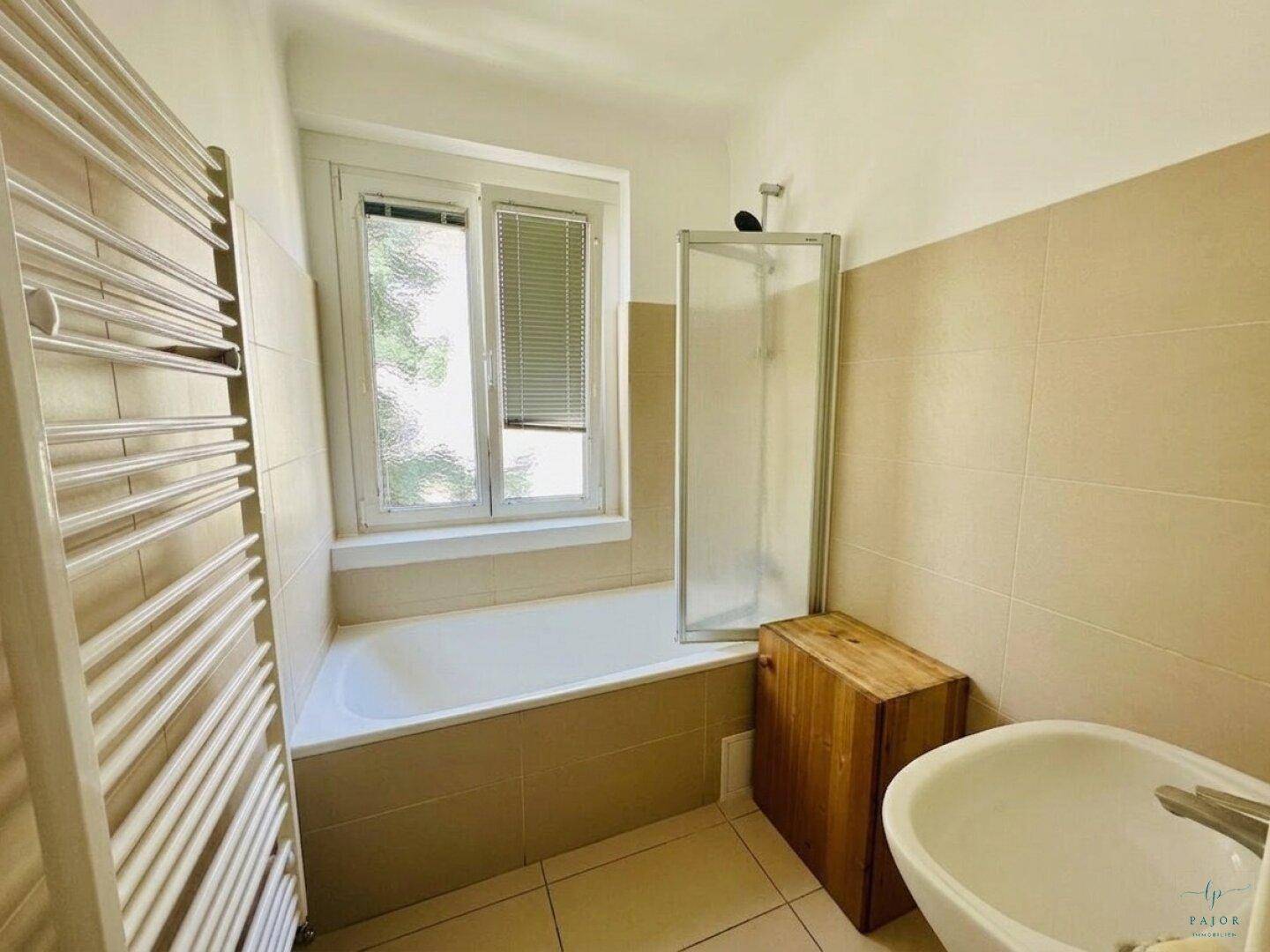 Badezimmer mit Badewanne, Duschabtrennung, beheiztem Handtuchhalter und Fenster, in neutralen Farbtönen.