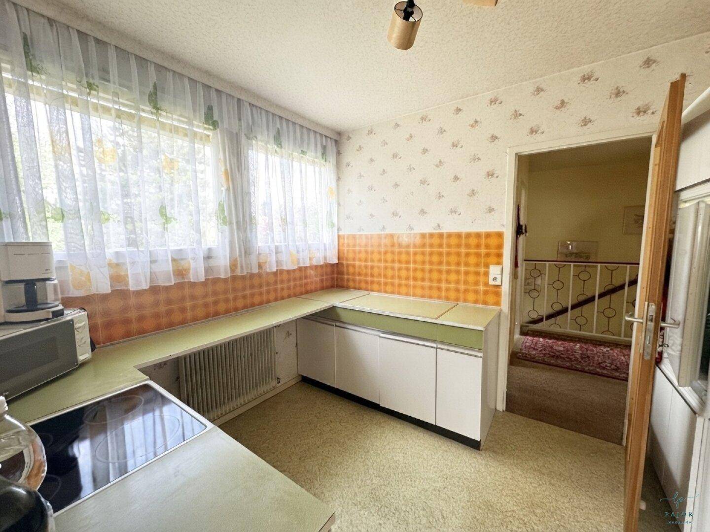 Küche im Retro-Stil mit grünen Arbeitsplatten, weißen Schränken und orangefarbenen Fliesen, hell durch großes Fenster.