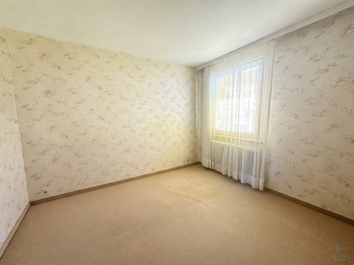 Helles, leeres Zimmer mit Teppichboden und großem Fenster, vielseitig nutzbar als Schlaf- oder Arbeitszimmer.