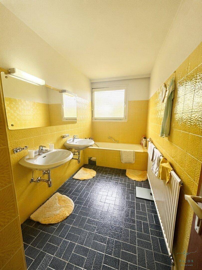 Badezimmer im 80er-Jahre-Stil mit gelben Wandfliesen, dunklem Boden und zwei Waschbecken sowie Badewanne.