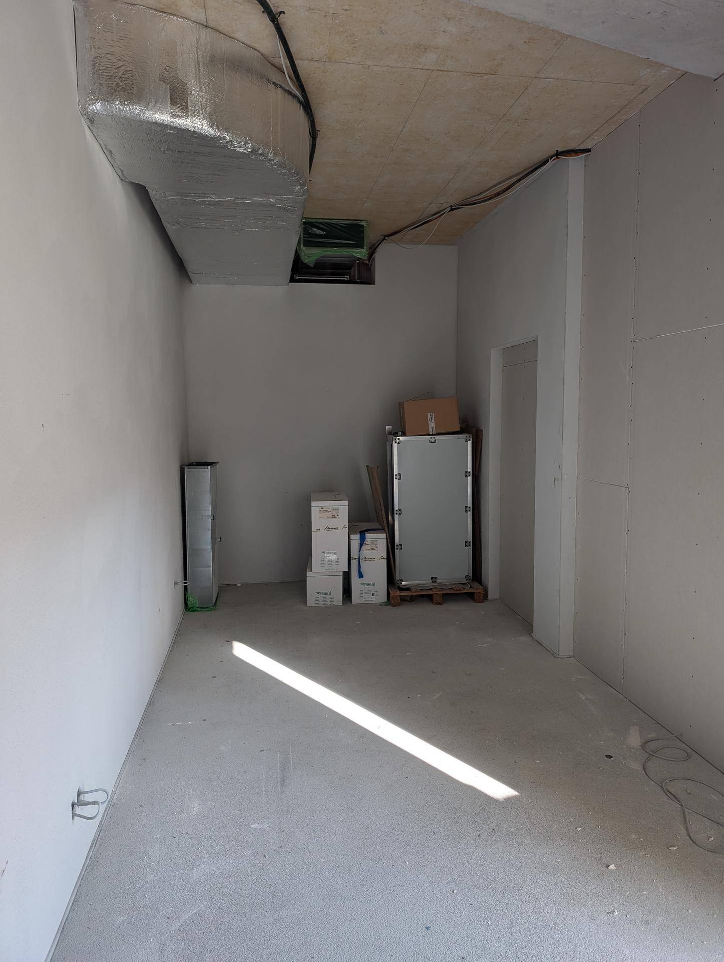 Unfertiger Innenraum mit Betonboden, sichtbaren Lüftungsrohren und einem Lichtstrahl.