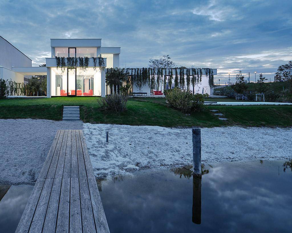 Moderne Villa am Seeufer in der Abenddämmerung mit Steg und beleuchteten Innenräumen.
