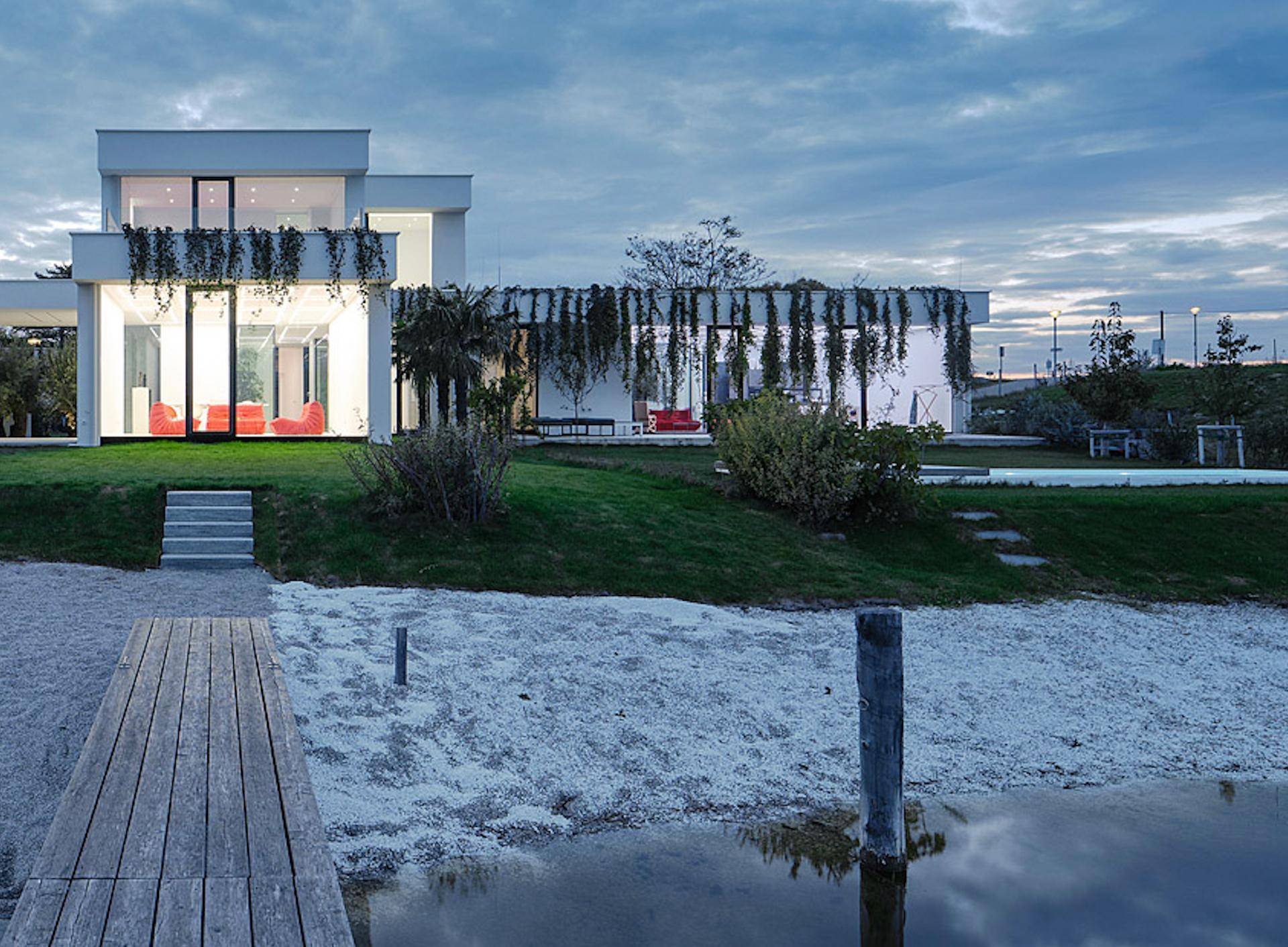 Moderne Architektur am Seeufer bei Dämmerung mit Steg und weitläufiger Grünfläche.