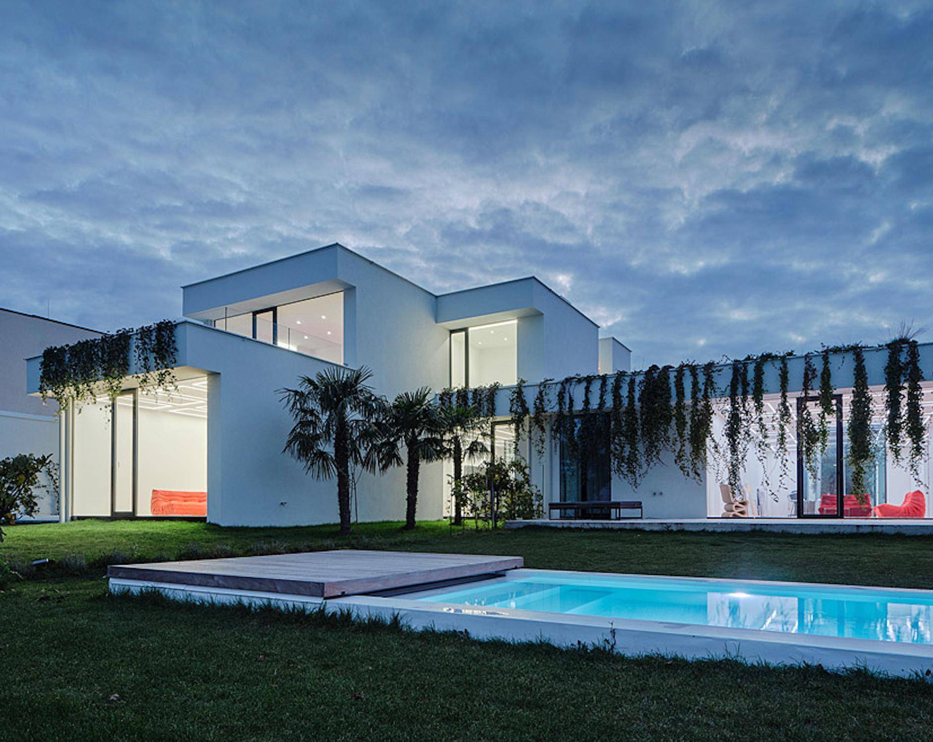 Moderne Villa bei Dämmerung mit beleuchtetem Pool und üppiger Begrünung.