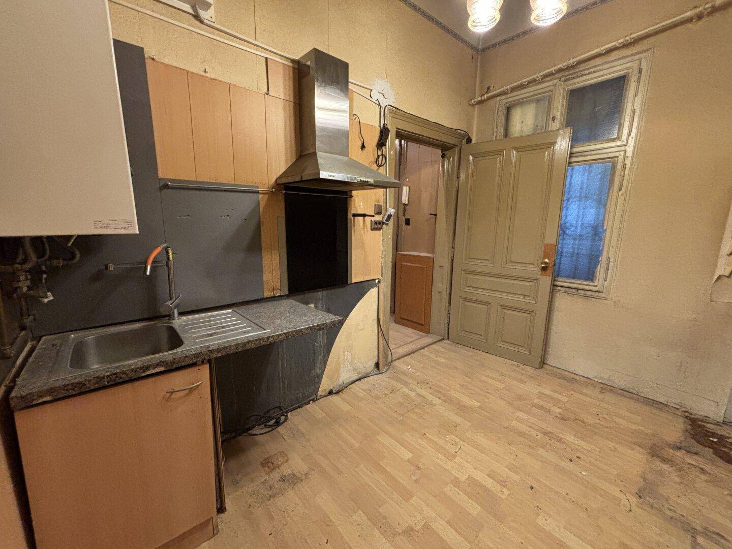 Abgenutzte Küche mit Spüle, Dunstabzugshaube und sichtbaren Installationsrohren.