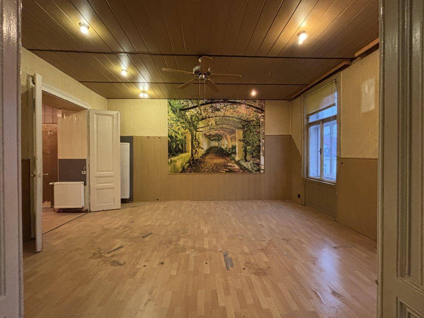 Geräumiges Wohnzimmer mit Holzdecke, Deckenventilator und abgenutztem Parkettboden.