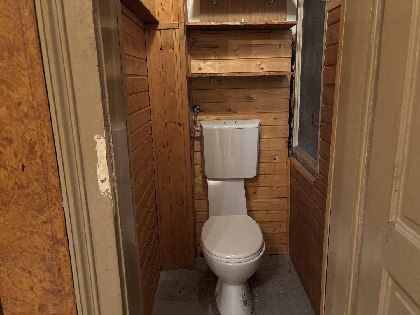 Kleine Toilette mit Holzverkleidung und Fenster, in einfachem Zustand.