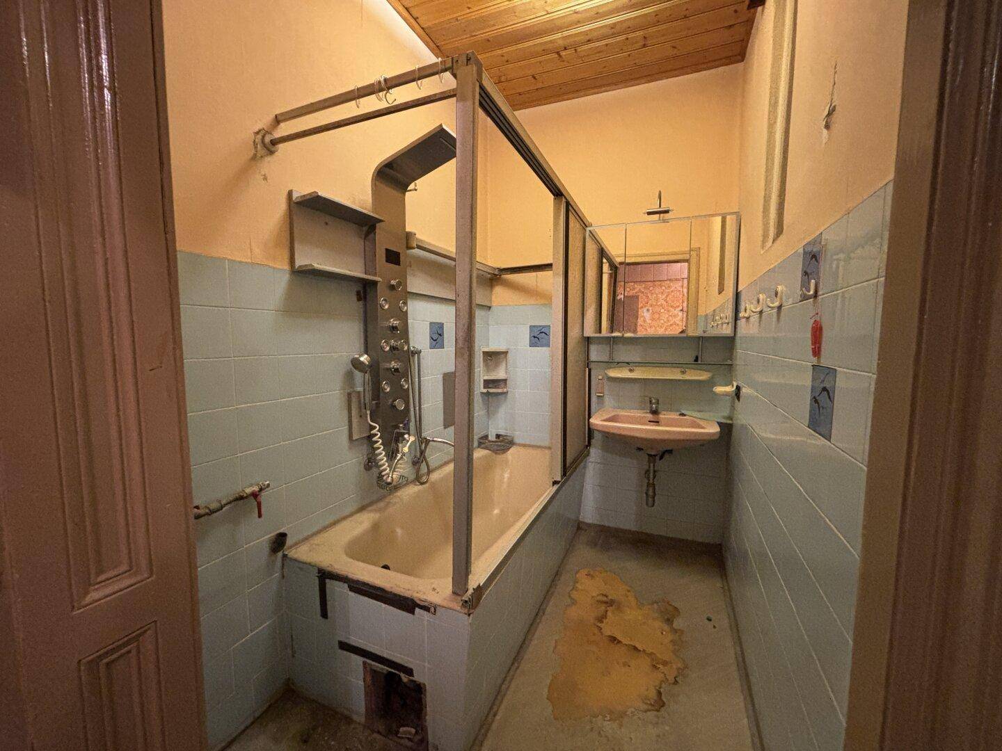 Altes Badezimmer mit Badewanne, blauen Fliesen und beschädigtem Boden.
