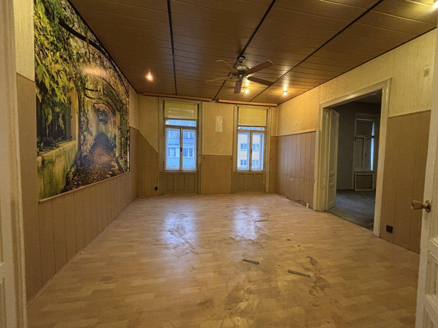 Geräumiges Wohnzimmer mit Deckenventilator, Holzboden und großen Fenstern.