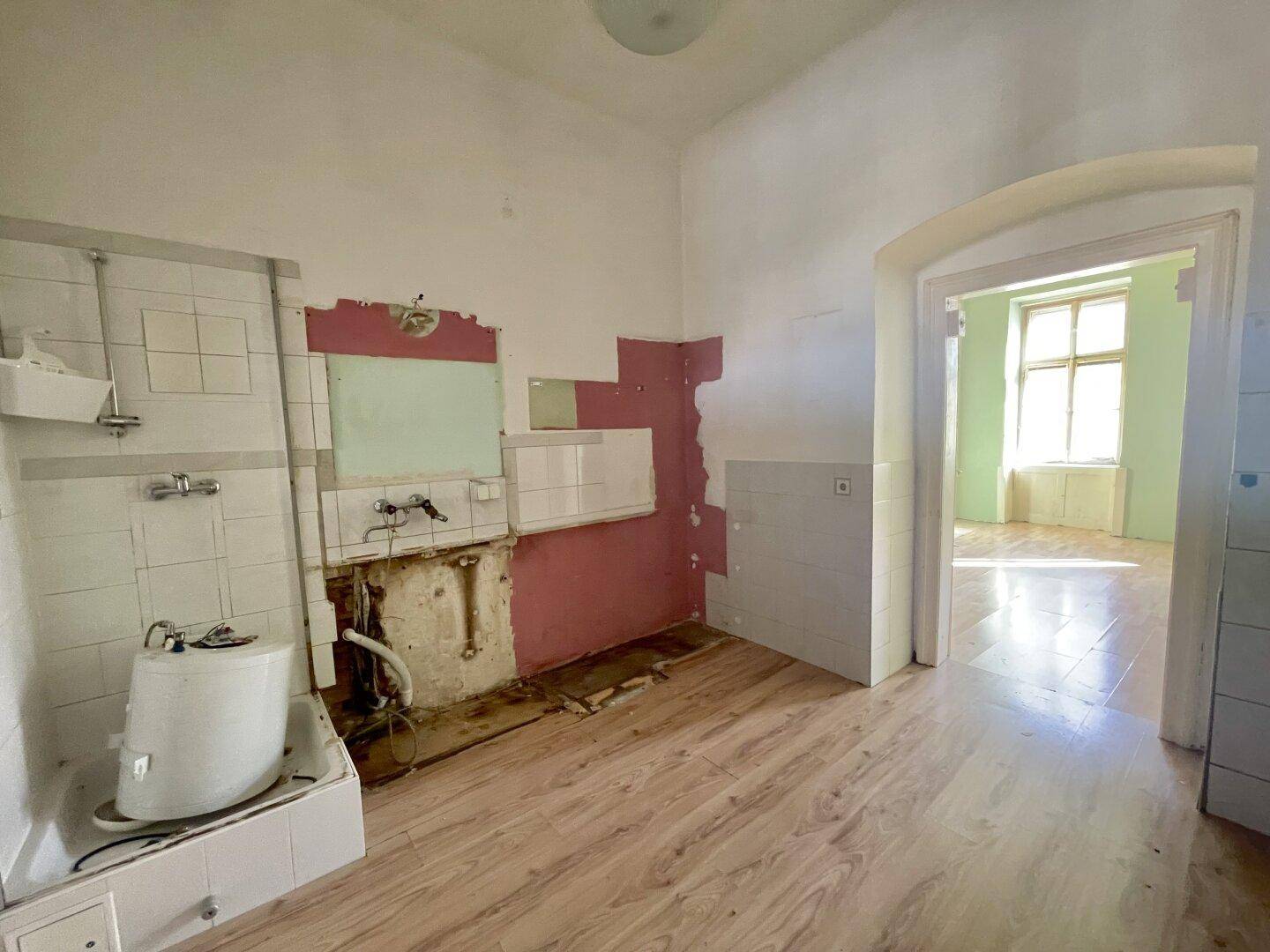 Renovierungsbedürftige Küche mit alten Fliesen, Holzboden und Blick in einen angrenzenden Raum.