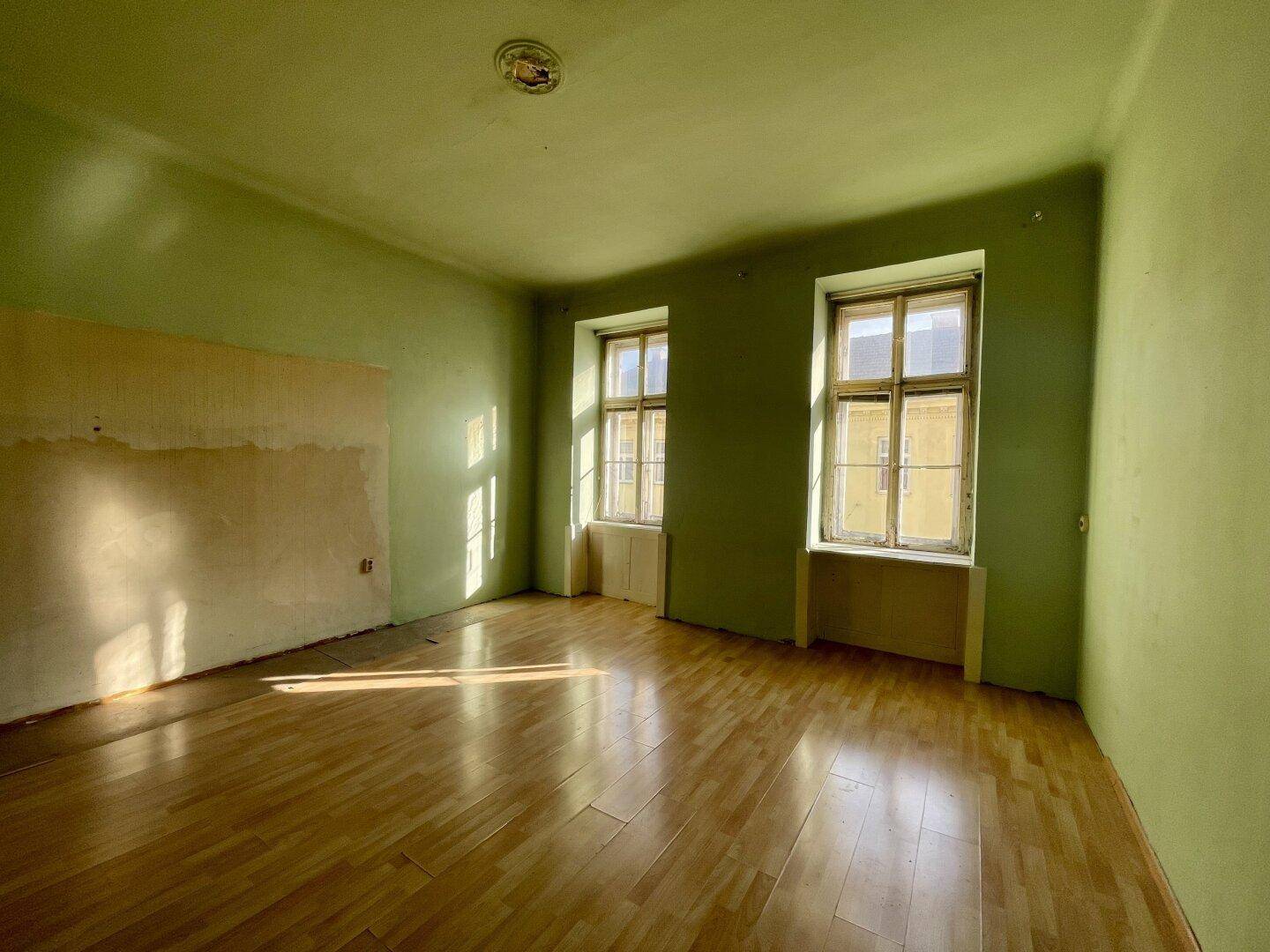 Geräumiges Zimmer mit hellgrünen Wänden, Holzboden und zwei großen Fenstern, die den Raum erhellen.