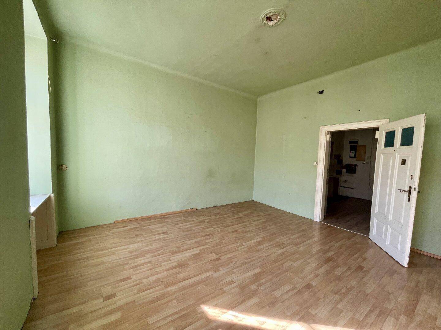 Leeres Wohnzimmer mit hellgrünen Wänden, Holzboden und einem Fenster, das viel Tageslicht hereinlässt.