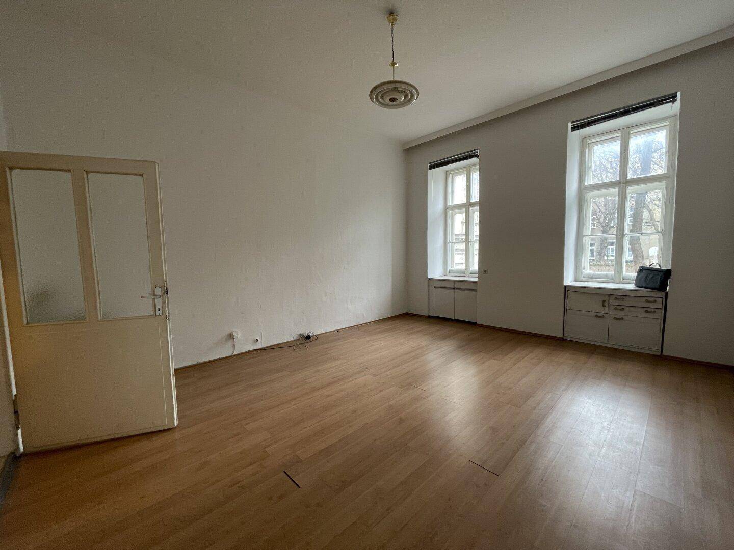 Heller, leerer Raum mit Holzboden, hohen Decken und zwei großen Fenstern, die viel Licht spenden.