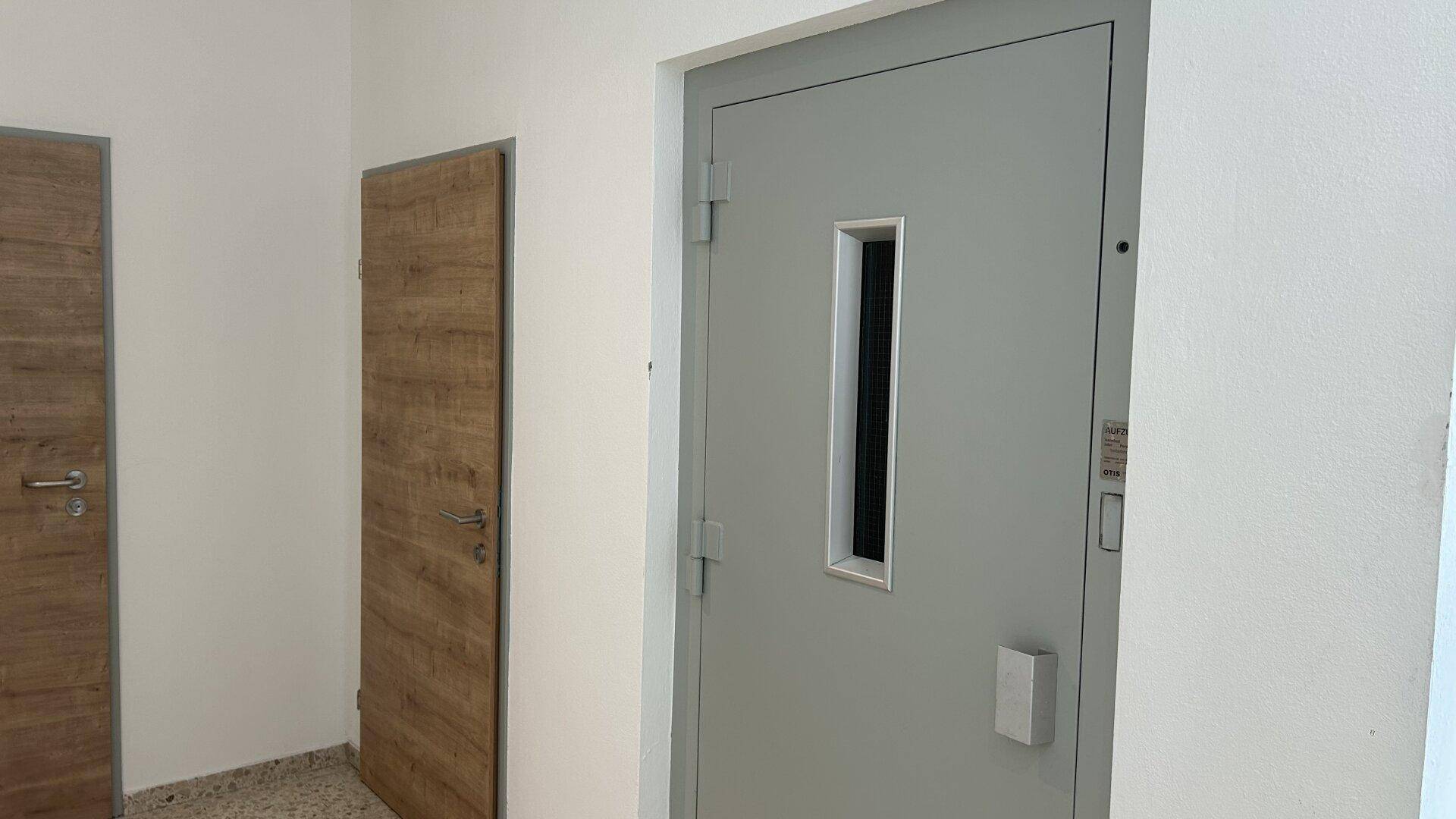 Heller Eingangsbereich mit Holztüren und einer grauen Aufzugstür, die den Zugang zu den Etagen ermöglicht.