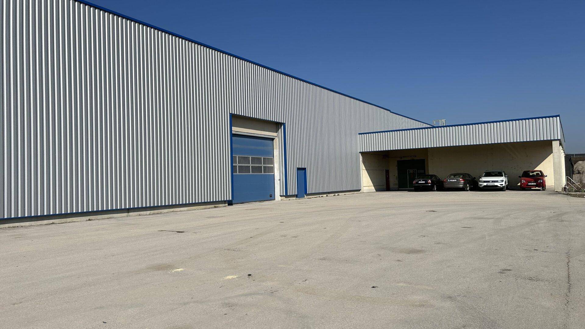 Moderne Fassade einer Lagerhalle mit Rolltor und Parkplätzen unter blauem Himmel.
