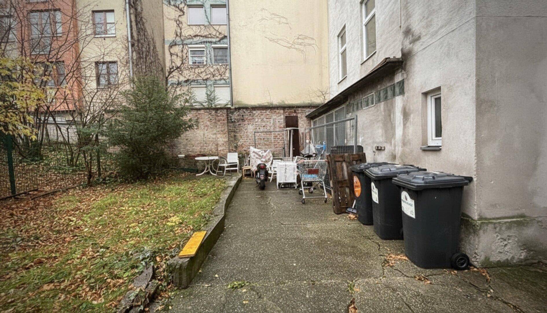 Hinterhofbereich mit Rasen, Mülltonnen und einem gepflasterten Weg.