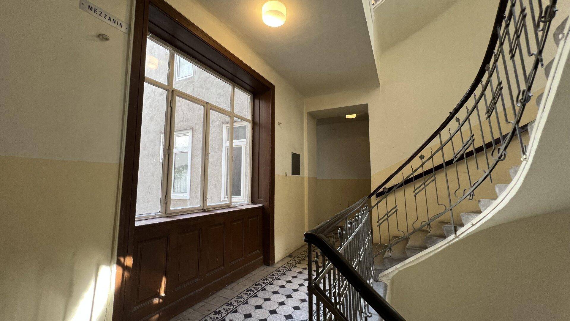 Helles Treppenhaus mit großem Fenster und traditionellem Fliesenboden, typisch für Altbauten.