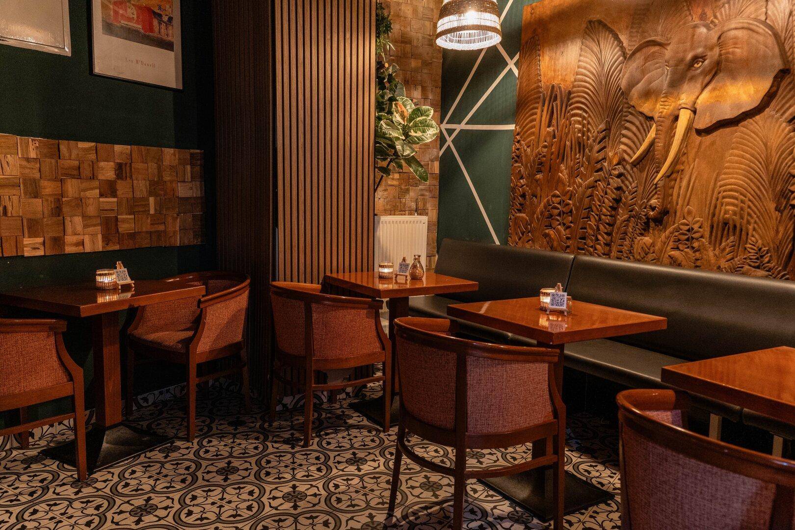 Gemütlicher Restaurantbereich mit Holztischen, gepolsterten Stühlen und einer kunstvollen Elefantenschnitzerei an der Wand.