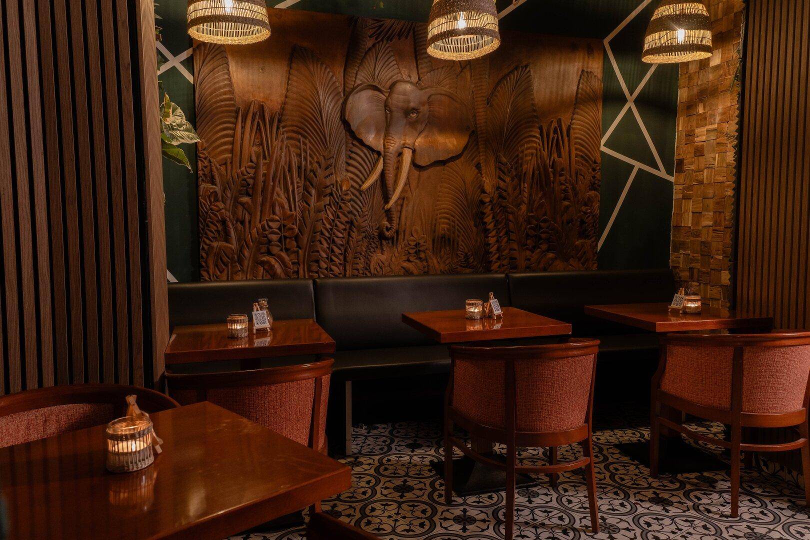 Ein stilvoller Restaurantbereich mit dunklen Sitzbänken, Holztischen und einer großen geschnitzten Elefantenwanddekoration.