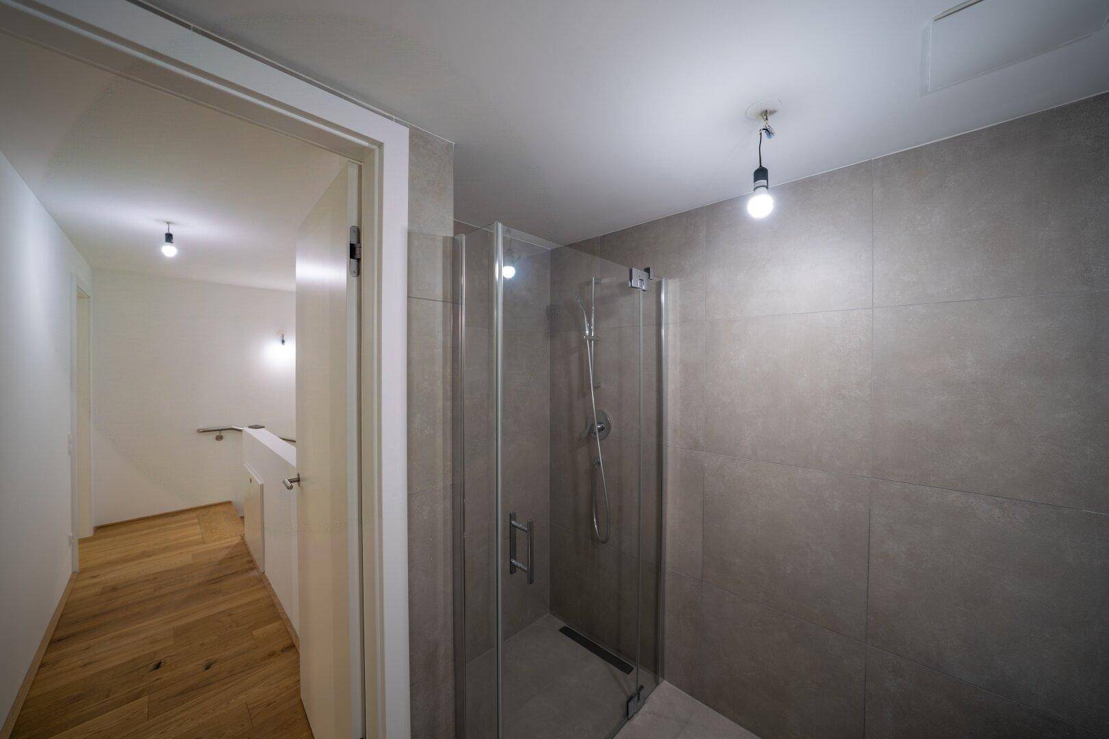Badezimmer mit moderner begehbarer Dusche und grauen Fliesen, offen zum Flur.