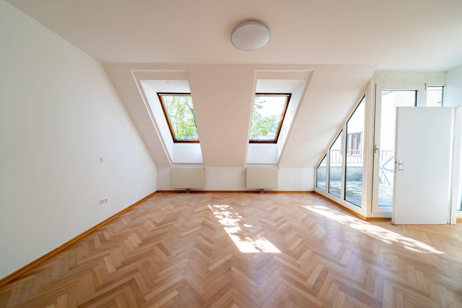 Heller Wohnraum mit Fischgrätparkett, Dachschrägen und großen Fenstern mit Zugang zur Terrasse.