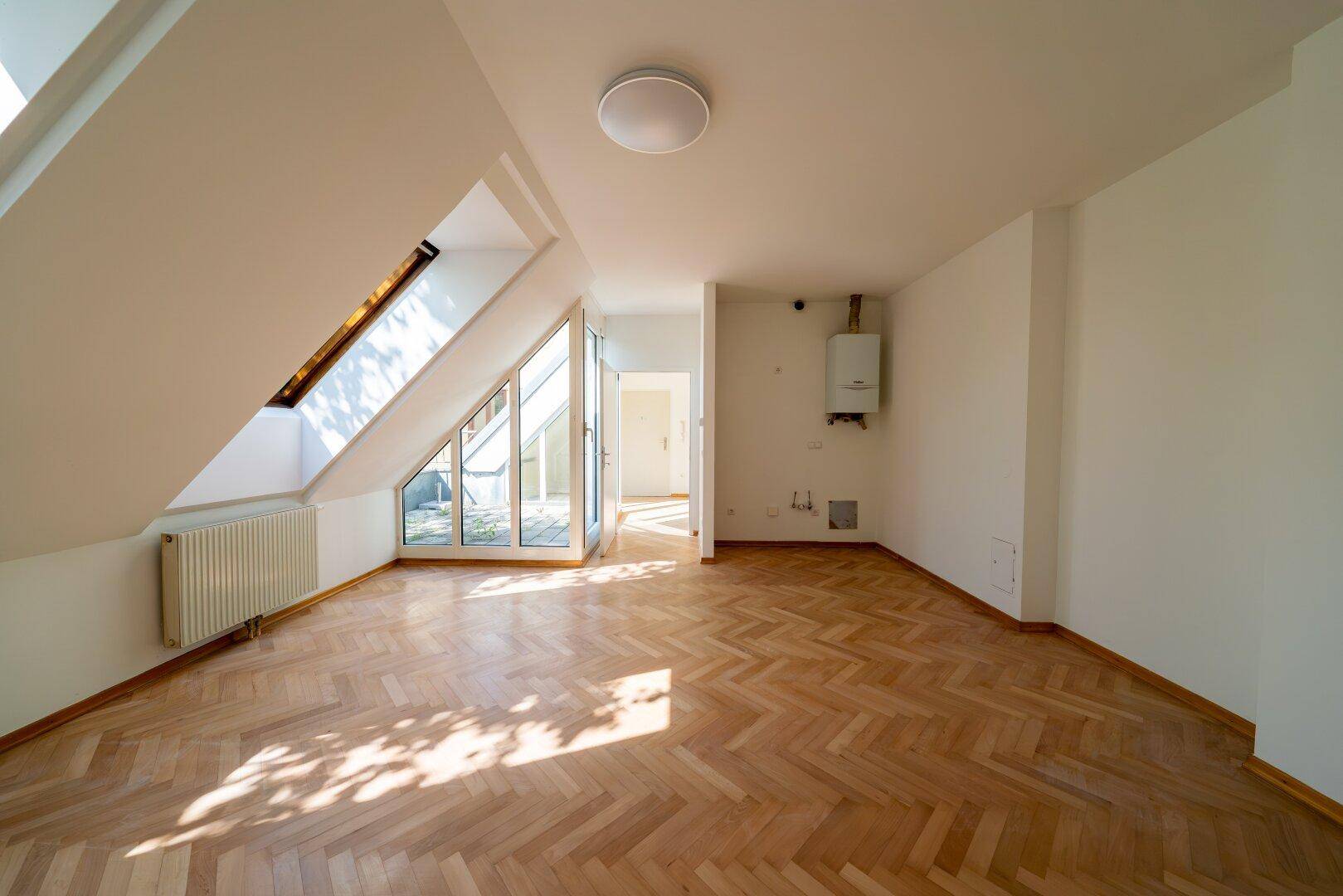 Geräumiger Wohnraum mit Fischgrätparkett, Dachschrägen und Zugang zu einer kleinen Terrasse.