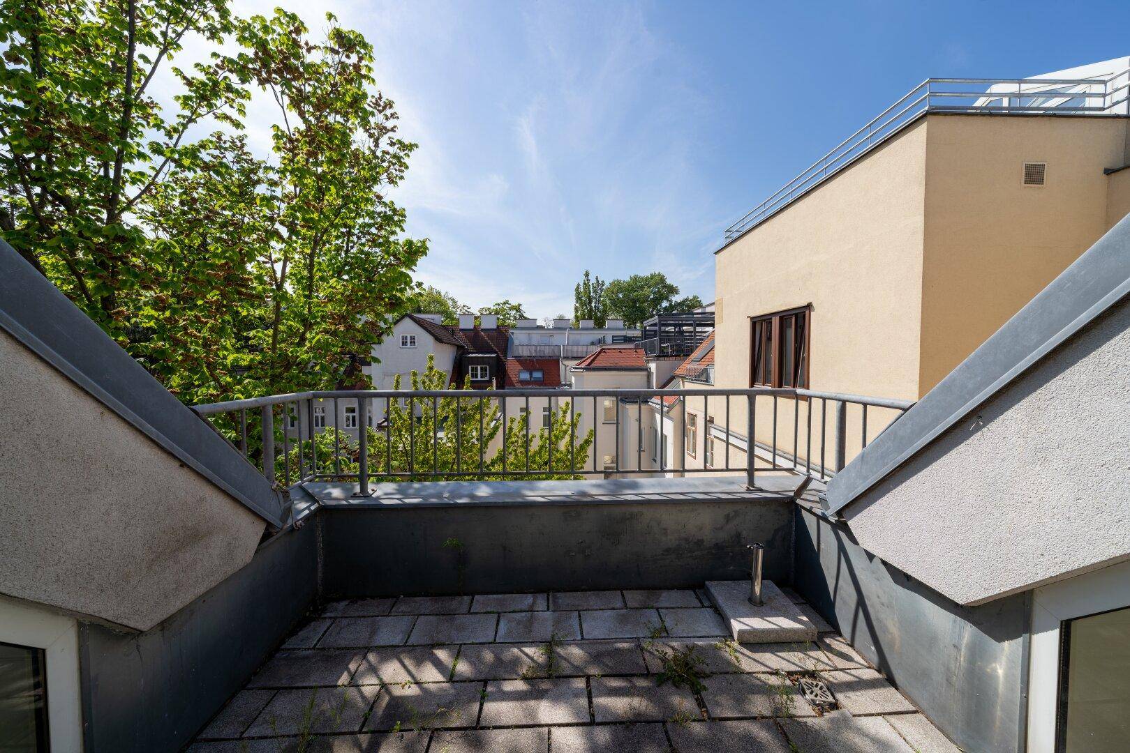 Kleine Dachterrasse mit Metallgeländer und Blick auf die umliegenden Gebäude und Bäume.