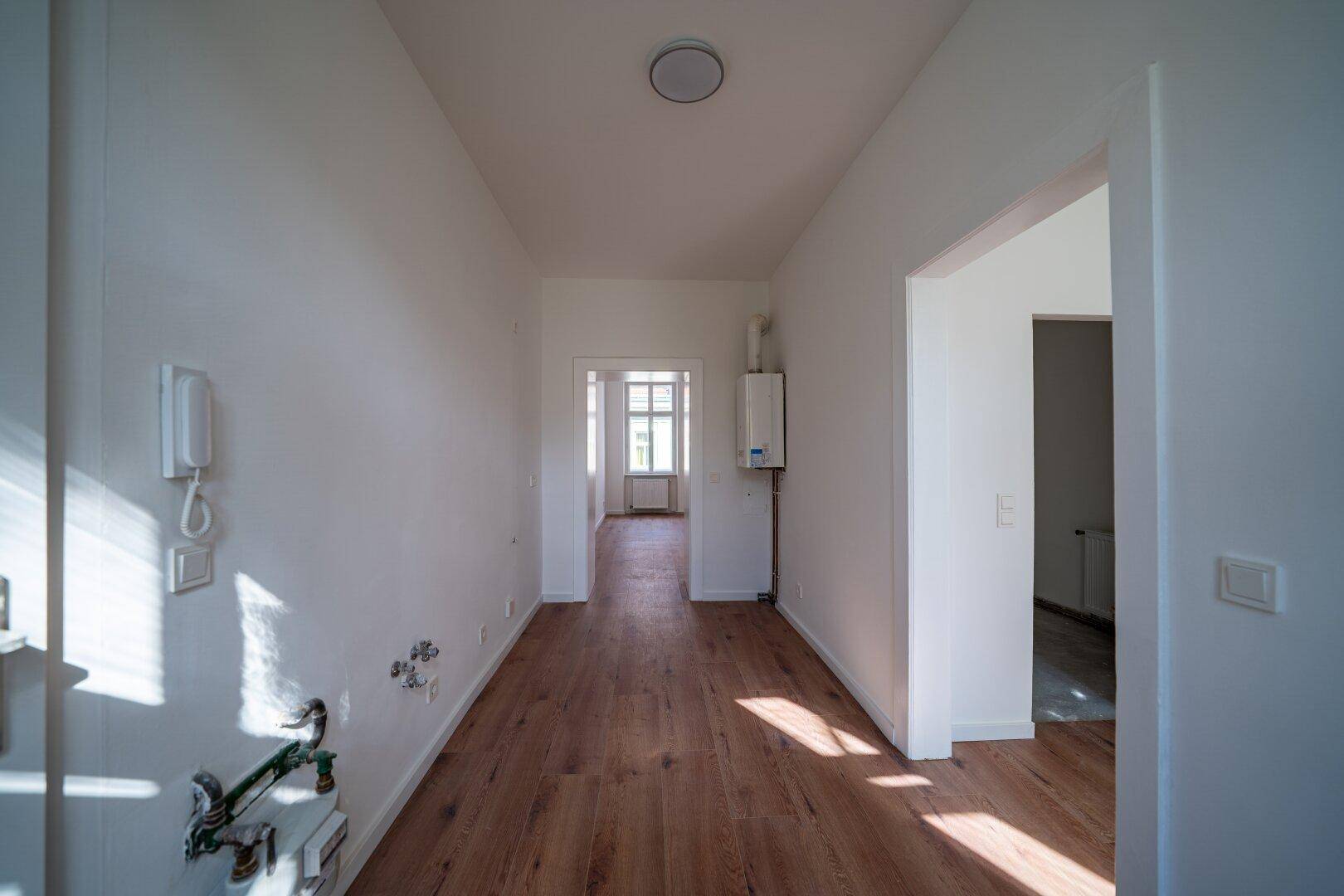 Heller Flur mit Holzboden und sichtbaren Installationen, der zu weiteren Räumen führt.