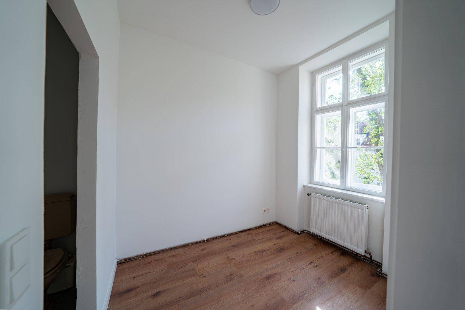 Kleines, helles Zimmer mit Holzboden, einem Fenster und Heizkörper, ideal als Schlaf- oder Arbeitszimmer.