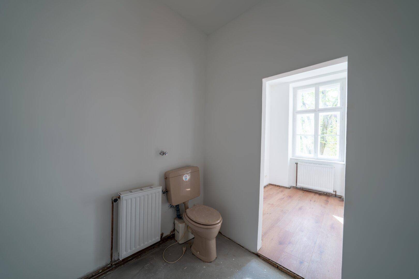 Einfaches WC mit beigefarbener Toilette und Heizkörper, angrenzend an einen weiteren Raum.