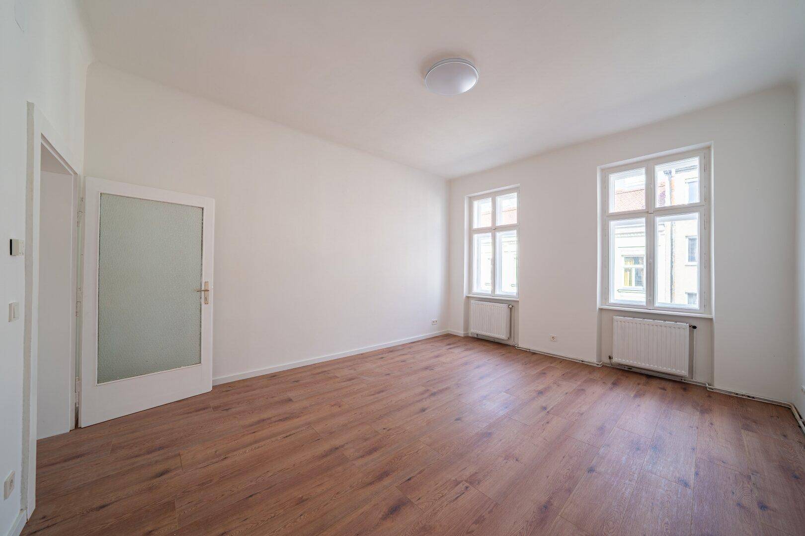 Geräumiges, helles Zimmer mit zwei Fenstern, Holzboden und weiß gestrichenen Wänden.