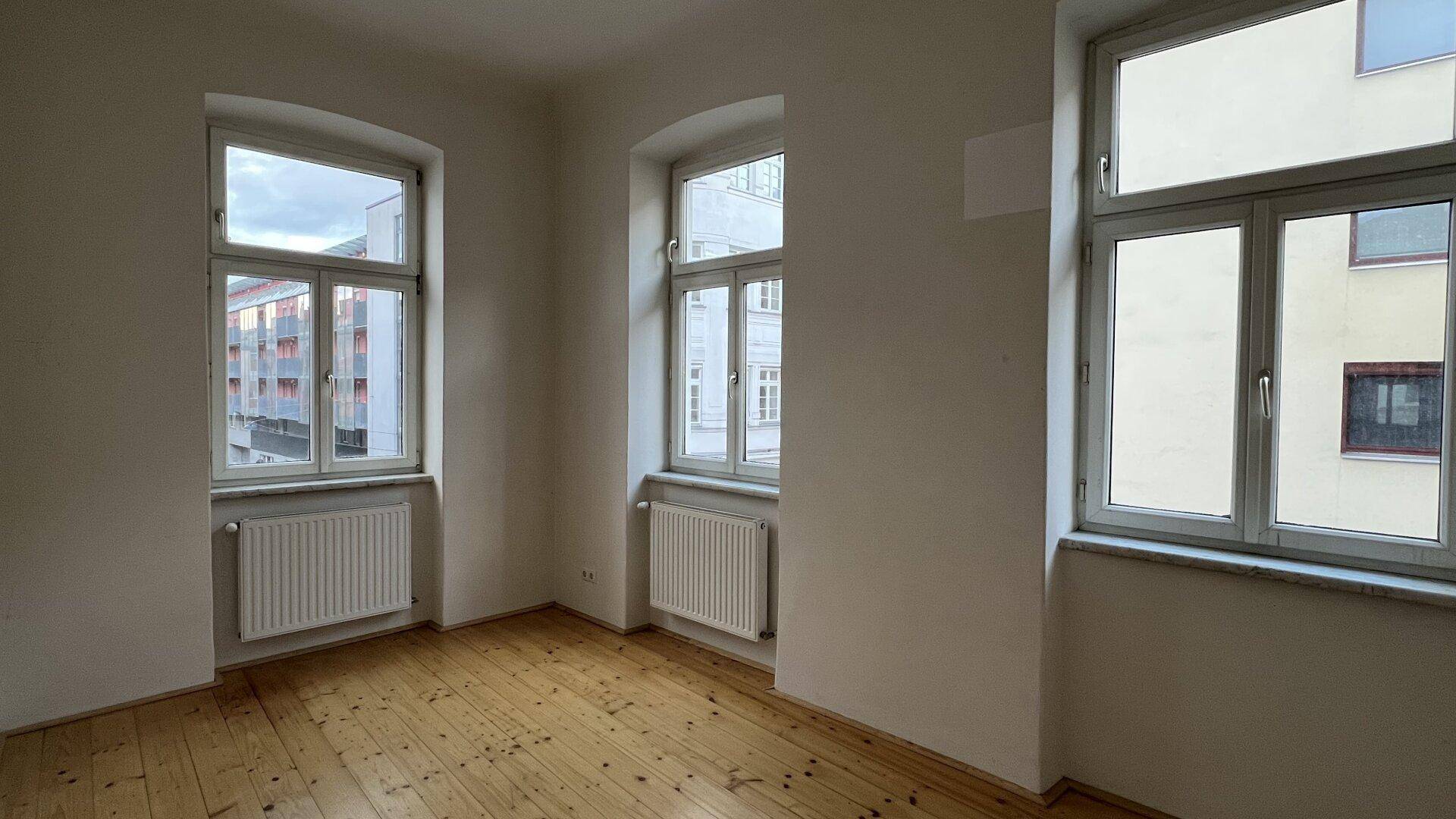 Leerer Raum mit hellem Holzplankenboden, weißen Wänden und drei großen Fenstern mit Heizkörpern.