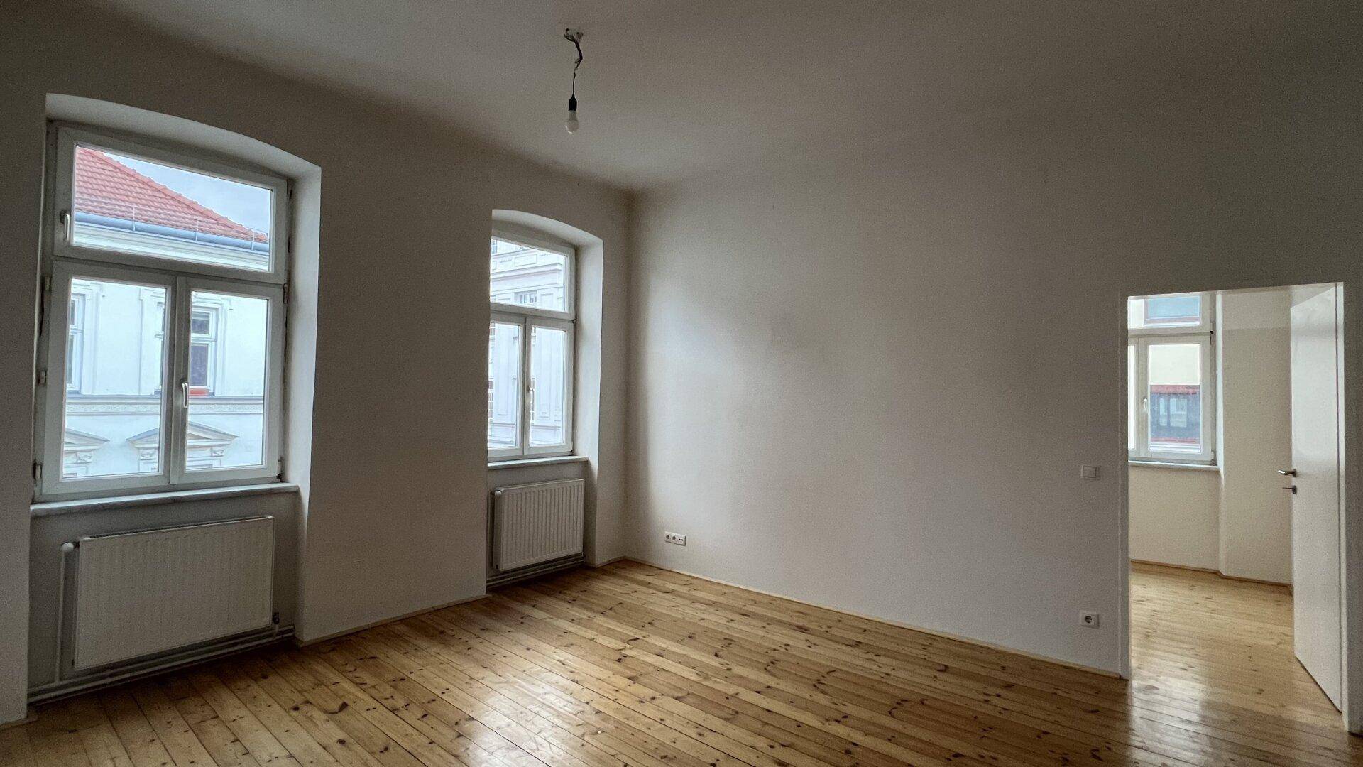 Geräumiger, leerer Raum mit Holzdielenboden, hohen Decken und zwei Fenstern, die viel Licht spenden.