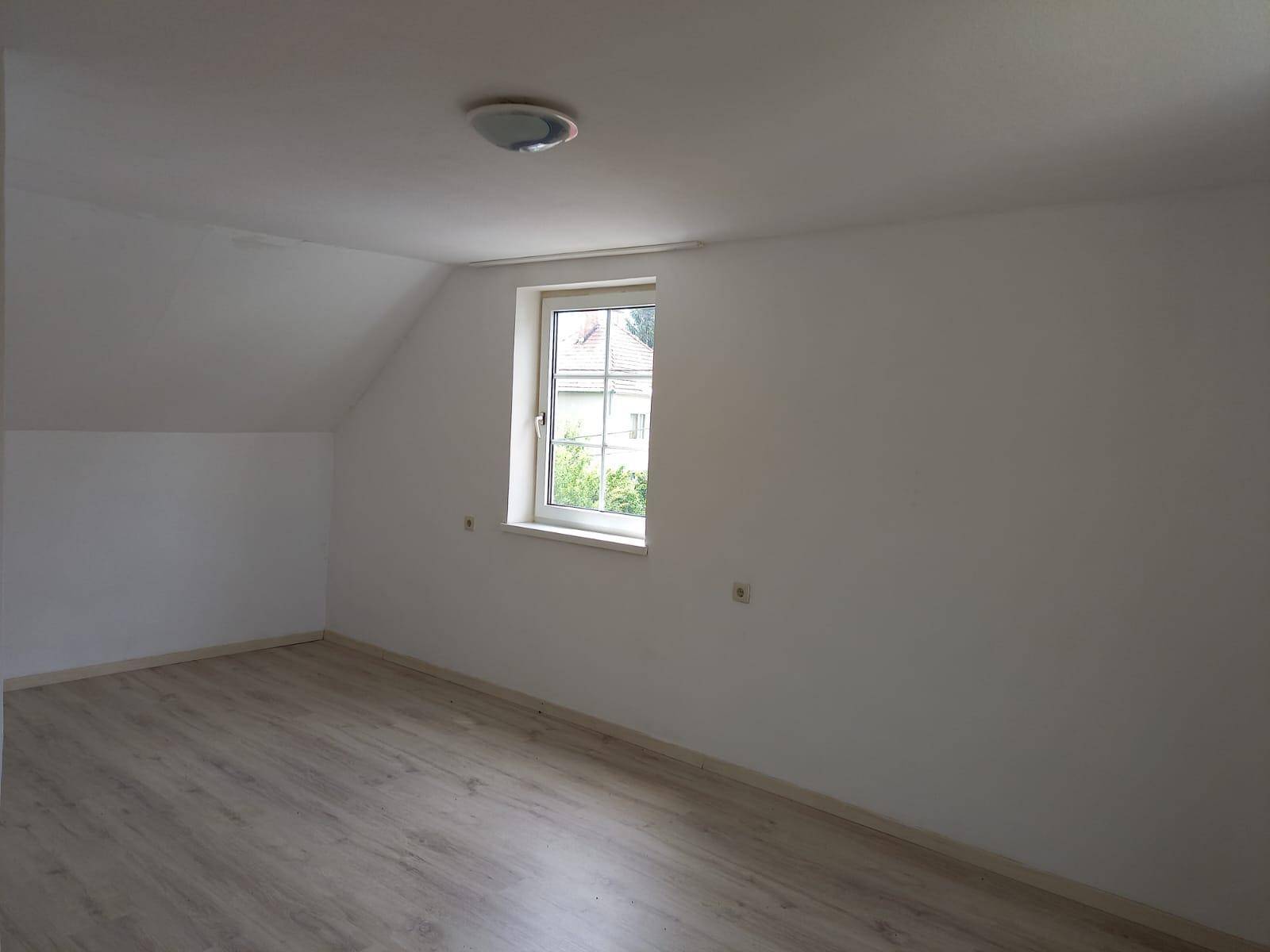 Dachgeschosszimmer mit schräger Decke, Laminatboden und einem Fenster mit Ausblick.
