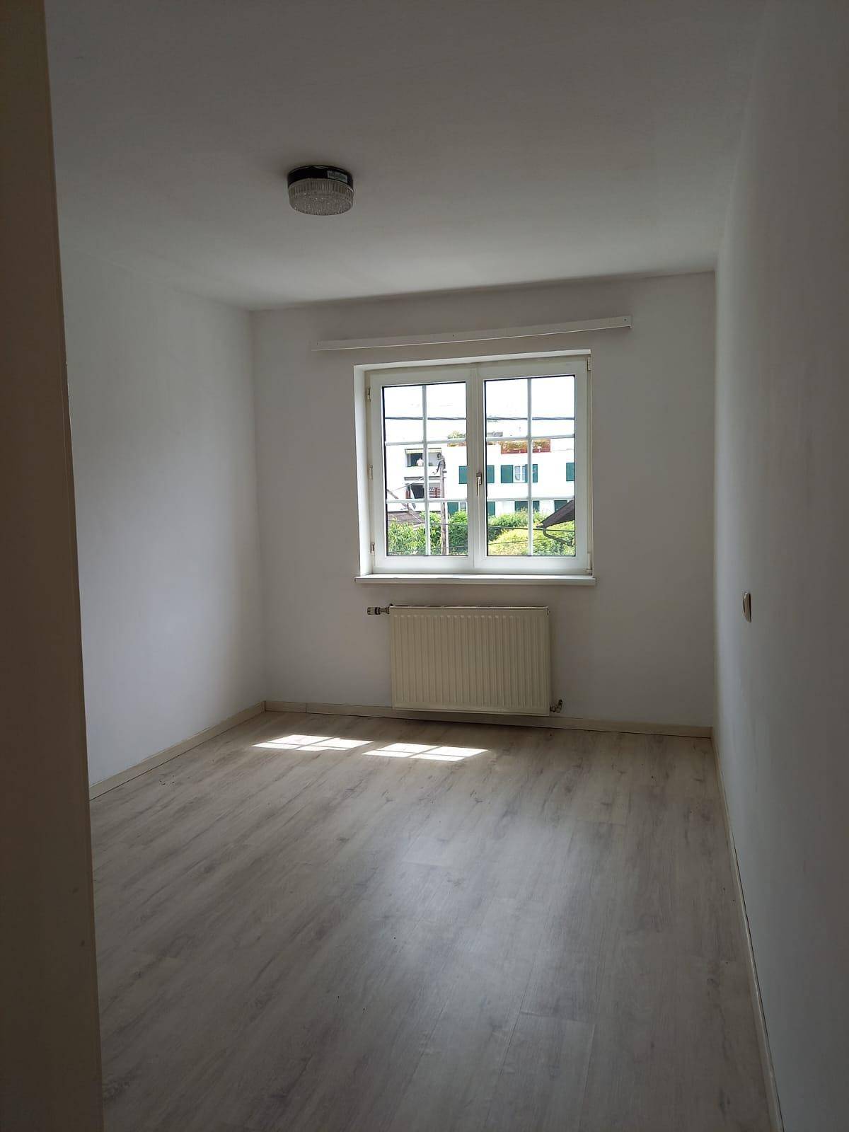 Helles Zimmer mit Laminatboden, Heizkörper und Blick ins Grüne durch das Fenster.