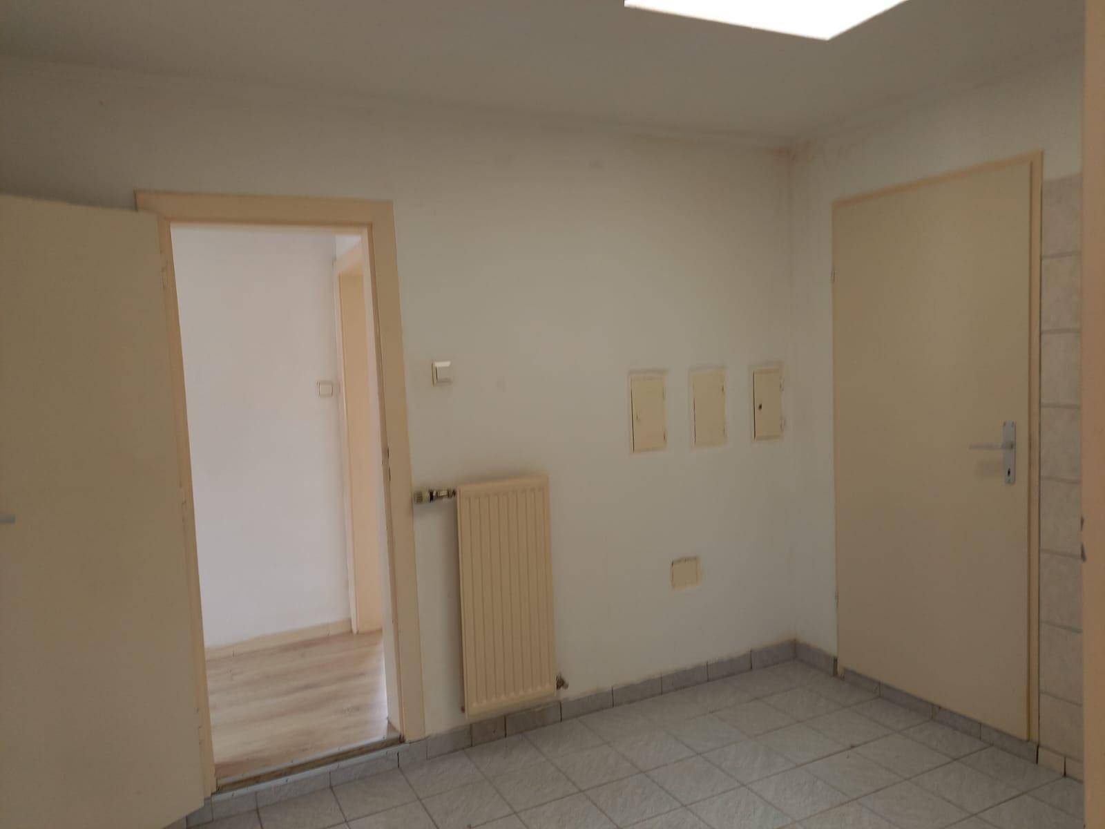 Einfacher Raum mit Fliesenboden, Heizkörper und mehreren Türen, möglicherweise ein Abstellraum.