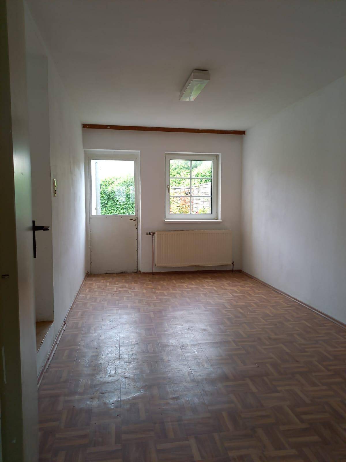 Helles Zimmer mit Fischgrätparkett, Fenster und Zugang zu einem Außenbereich.
