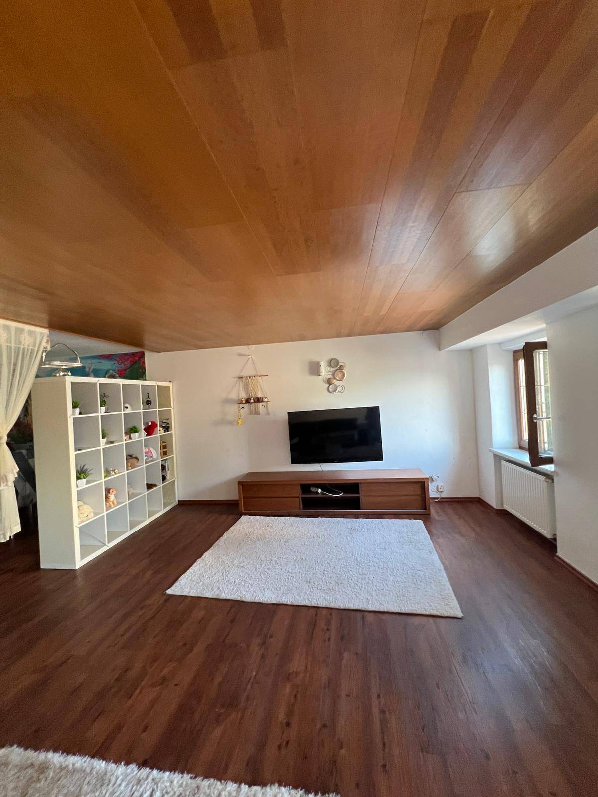 Heller Wohnbereich mit dunklem Laminatboden, Holzdecke und einem weißen Regal als Raumteiler.