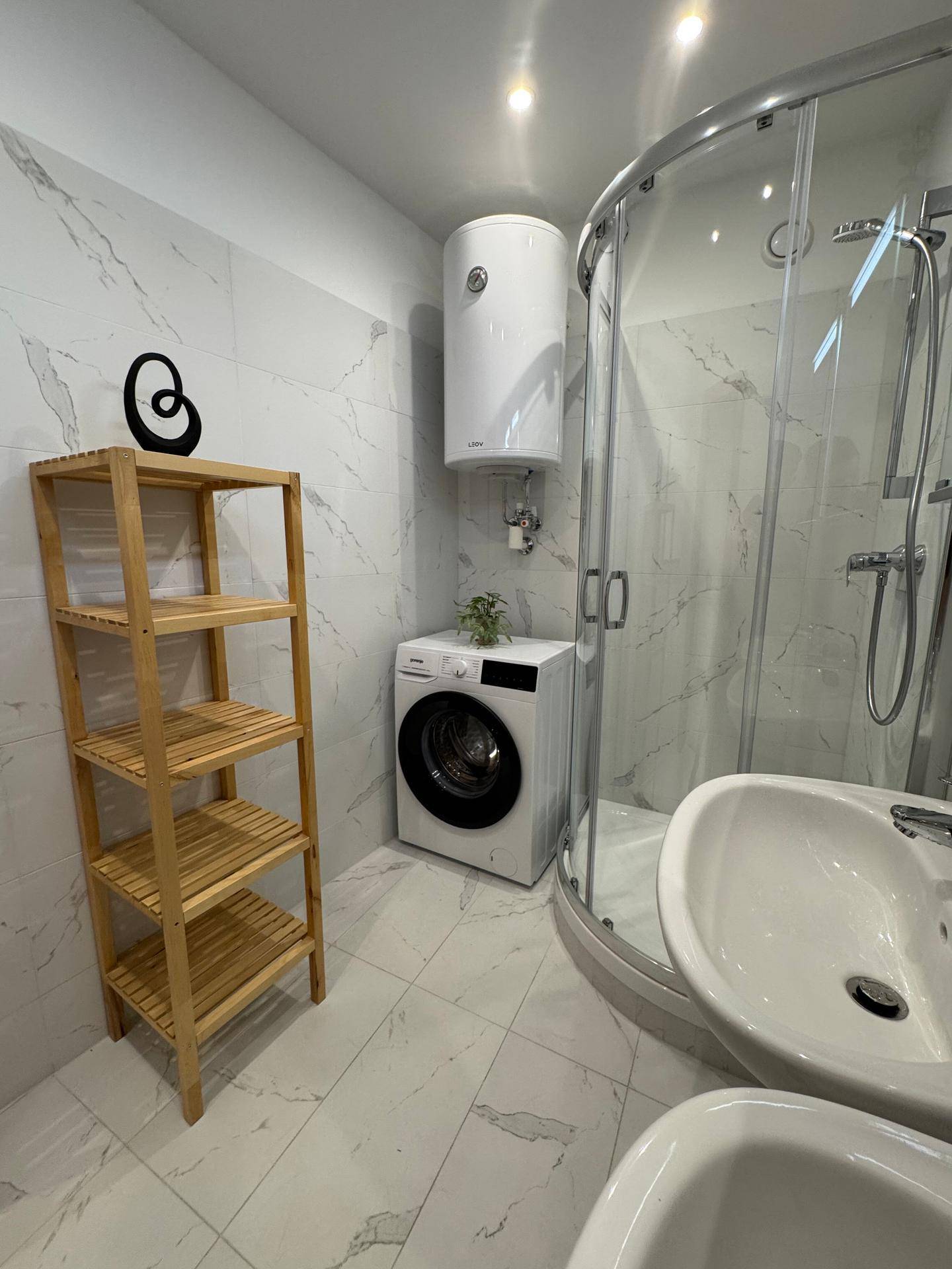 Helles Badezimmer mit Duschkabine, Waschmaschine und einem praktischen Holzregal.