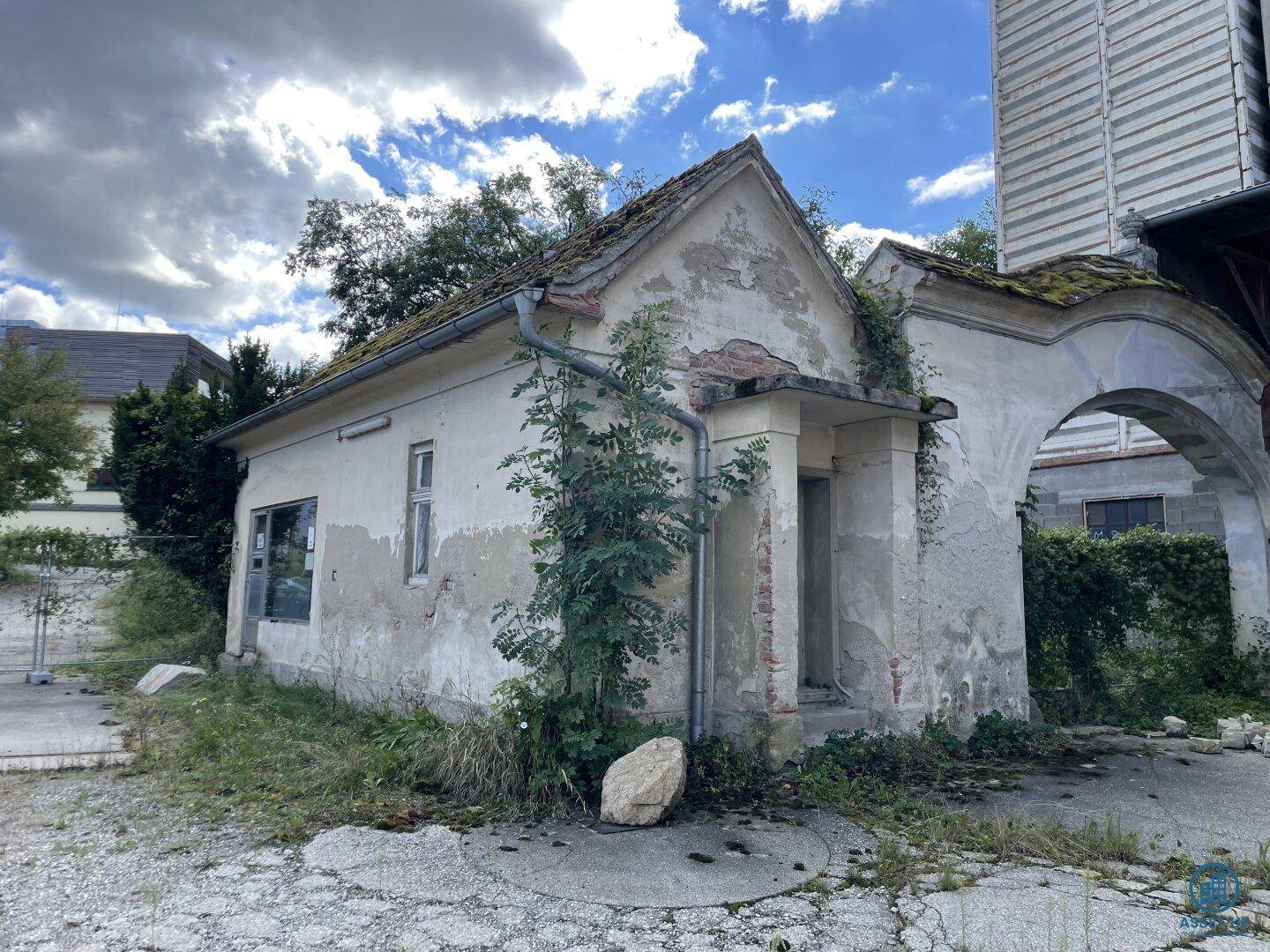 Verfallenes kleines Gebäude mit bröckelndem Putz und überwucherten Mauern neben einem alten Torbogen.