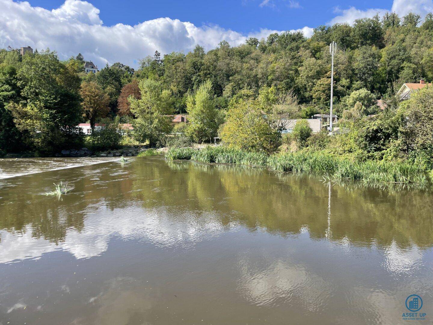Breiter Flusslauf umgeben von üppiger grüner Vegetation und bewaldeten Hügeln unter blauem Himmel.